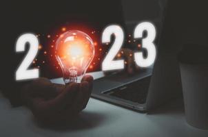 mano de negocios con texto de luz de bombilla 2023 y computadora portátil en el escritorio.concepto de plan de crecimiento de innovación de idea. foto