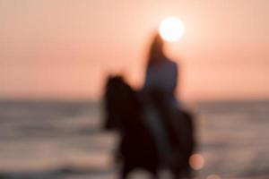 Blurred photo. Woman in summer clothes enjoys riding a horse on a beautiful sandy beach at sunset. Selective focus photo