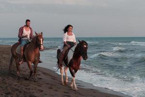 una joven pareja amorosa vestida de verano montando a caballo en una playa de arena al atardecer. mar y puesta de sol de fondo. enfoque selectivo foto