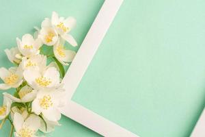 Beautiful white flowers and blank frame on pastel mint background. Top view, copy space. photo