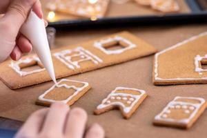 mujer joven está decorando galletas de casa de pan de jengibre de navidad en casa con cobertura de glaseado en bolsa de hielo, primer plano, estilo de vida. foto