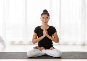 Asian women doing yoga meditating exercise at home, sitting in Lotus pose or Padmasana with raised hands namaste in white bedroom. Working out wearing black shirt and white pants. Healthcare concept. photo