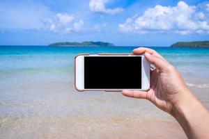 Tourist using phone at the beach with the sea, hand holding white mobile smart phone smartphone, travel working concept, blurry background, close up. photo