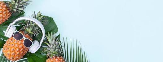 Funny pineapple wearing white headphone, concept of listening music, isolated on blue background with tropical palm leaves, top view, flat lay design. photo