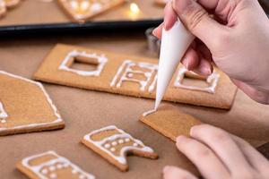 mujer joven está decorando galletas de casa de pan de jengibre de navidad en casa con cobertura de glaseado en bolsa de hielo, primer plano, estilo de vida. foto