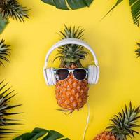 Funny pineapple wearing white headphone, listen music, isolated on yellow background with tropical palm leaves, top view, flat lay design concept. photo