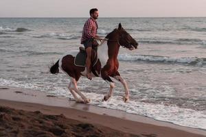 un hombre moderno con ropa de verano disfruta montando a caballo en una hermosa playa de arena al atardecer. enfoque selectivo foto