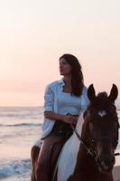 Woman in summer clothes enjoys riding a horse on a beautiful sandy beach at sunset. Selective focus photo