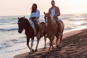 una pareja amorosa vestida de verano montando a caballo en una playa de arena al atardecer. mar y puesta de sol de fondo. enfoque selectivo foto