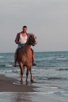 A modern man in summer clothes enjoys riding a horse on a beautiful sandy beach at sunset. Selective focus photo