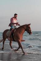 A modern man in summer clothes enjoys riding a horse on a beautiful sandy beach at sunset. Selective focus photo