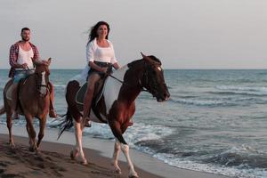 una pareja amorosa vestida de verano montando a caballo en una playa de arena al atardecer. mar y puesta de sol de fondo. enfoque selectivo foto