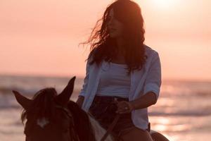 Woman in summer clothes enjoys riding a horse on a beautiful sandy beach at sunset. Selective focus photo