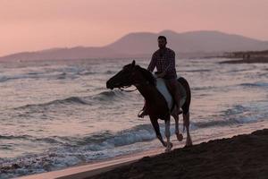 A modern man in summer clothes enjoys riding a horse on a beautiful sandy beach at sunset. Selective focus photo