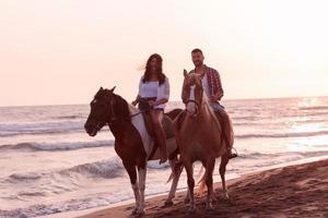 una pareja amorosa vestida de verano montando a caballo en una playa de arena al atardecer. mar y puesta de sol de fondo. enfoque selectivo foto