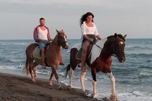 una pareja amorosa vestida de verano montando a caballo en una playa de arena al atardecer. mar y puesta de sol de fondo. enfoque selectivo foto