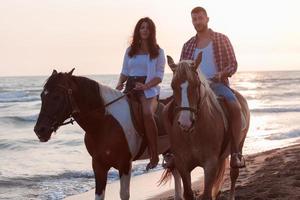 una pareja amorosa vestida de verano montando a caballo en una playa de arena al atardecer. mar y puesta de sol de fondo. enfoque selectivo foto