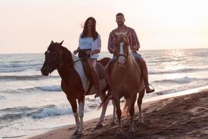 una pareja amorosa vestida de verano montando a caballo en una playa de arena al atardecer. mar y puesta de sol de fondo. enfoque selectivo foto