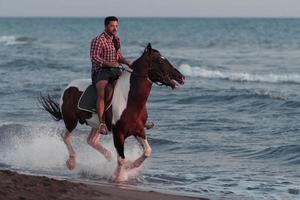 un hombre moderno con ropa de verano disfruta montando a caballo en una hermosa playa de arena al atardecer. enfoque selectivo foto