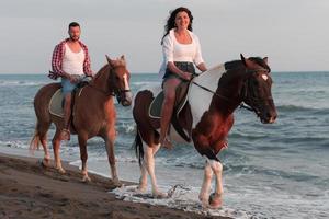 una pareja amorosa vestida de verano montando a caballo en una playa de arena al atardecer. mar y puesta de sol de fondo. enfoque selectivo foto