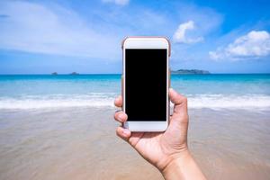 Tourist using phone at the beach with the sea, hand holding white mobile smart phone smartphone, travel working concept, blurry background, close up. photo