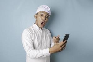 hombre balinés sorprendido con udeng o diadema tradicional y camisa blanca sosteniendo su teléfono inteligente, aislado de fondo azul foto