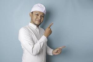 Excited Balinese man wearing udeng or traditional headband and white shirt pointing at the copy space beside him, isolated by blue background photo