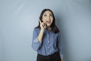 Excited Asian woman wearing blue shirt pointing at the copy space upwards, isolated by blue background photo