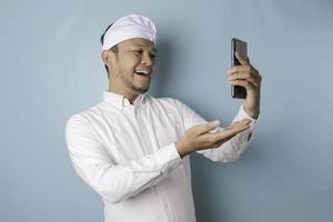 un retrato de un hombre balinés feliz sonríe y sostiene su teléfono inteligente usando udeng o una diadema tradicional y un pantalones blanco aislado por un fondo azul foto