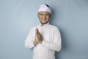 sonriente joven balinés con udeng o diadema tradicional y camisa blanca haciendo gestos de saludo o namaste aislado sobre fondo azul foto