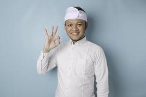 Excited Balinese man wearing udeng or traditional headband and white shirt giving an OK hand gesture isolated by a blue background photo