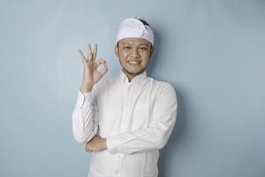 emocionado hombre balinés con udeng o diadema tradicional y camisa blanca dando un gesto de mano bien aislado por un fondo azul foto