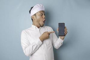 Surprised Balinese man wearing udeng or traditional headband and white shirt holding his smartphone, isolated by blue background photo