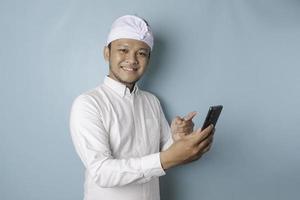 A portrait of a happy Balinese man is smiling and holding his smartphone wearing udeng or traditional headband and white shirt isolated by a blue background photo