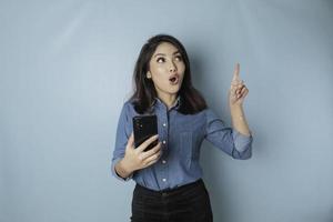 Surprised Asian woman wearing blue shirt pointing at the copy space upside her while holding a smartphone, isolated by blue background photo