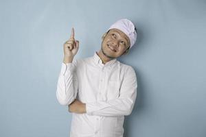 Excited Balinese man wearing udeng or traditional headband and white shirt pointing at the copy space upside him, isolated by blue background photo