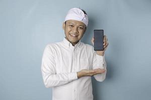 Excited Balinese man wearing traditional headband or udeng and white shirt pointing at the copy space on his smartphone, isolated by blue background photo