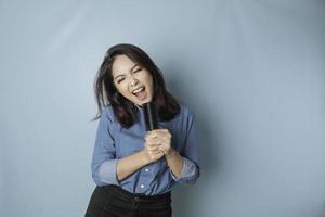 Portrait of carefree Asian woman, having fun karaoke, singing in microphone while standing over blue background photo