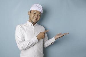 Excited Balinese man wearing udeng or traditional headband and white shirt pointing at the copy space beside him, isolated by blue background photo