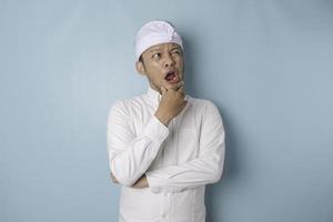 Portrait of a thoughtful young Balinese man wearing udeng or traditional headband and white shirt looking aside while his finger on his chin isolated over blue background photo