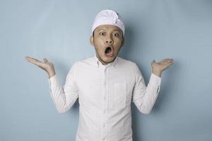 Surprised Balinese man wearing udeng or traditional headband and white shirt, isolated by blue background