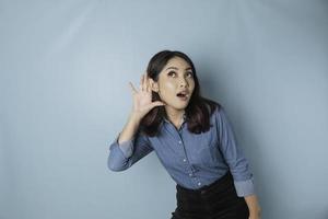 asombrada y curiosa mujer joven con una camisa azul tratando de oírte escuchar atentamente aislada en un retrato de estudio de fondo azul turquesa. foto