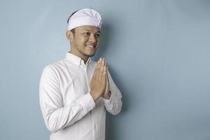 Smiling young Balinese man wearing udeng or traditional headband and white shirt gesturing greeting or namaste isolated over blue background photo