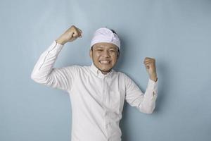 un joven balinés con una expresión feliz y exitosa que usa udeng o diadema tradicional y pantalones blanco aislado de fondo azul foto