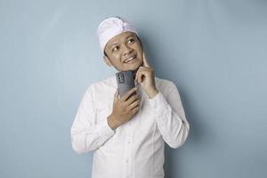 retrato de un joven balinés pensativo que usa udeng o diadema tradicional y camisa blanca mirando a un lado mientras sostiene un teléfono inteligente aislado sobre fondo azul foto