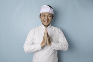 sonriente joven balinés con udeng o diadema tradicional y camisa blanca haciendo gestos de saludo o namaste aislado sobre fondo azul foto