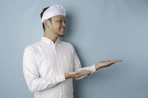 Excited Balinese man wearing udeng or traditional headband and white shirt pointing at the copy space beside him, isolated by blue background photo