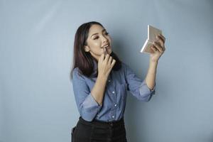 A portrait of a beautiful young Asian woman applying lipstick on her lips, isolated by a blue background photo