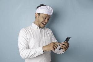 A portrait of a happy Balinese man is smiling and holding his smartphone wearing udeng or traditional headband and white shirt isolated by a blue background photo