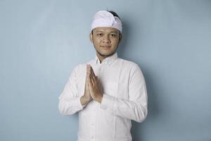 sonriente joven balinés con udeng o diadema tradicional y camisa blanca haciendo gestos de saludo o namaste aislado sobre fondo azul foto